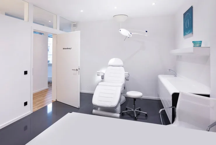 Heller Behandlungsraum bei California Skin München mit weißer Liege, Behandlungsleuchte und Waschtisch.